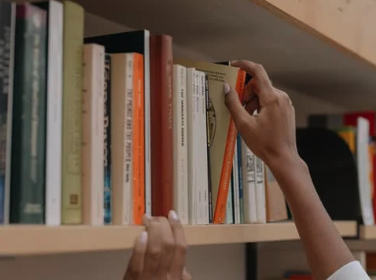 Close-up of a person’s hands reaching for a book on a wooden bookshelf filled with books of various sizes and colors. The person appears to be selecting an orange book from the middle of the shelf.